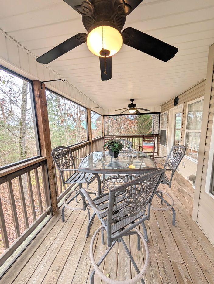 Screened back porch at The Smoky Nest with a glass-top table, four tall patio chairs, and ceiling fans, overlooking trees